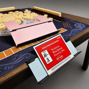 May include: A complete Mahjong set on a wooden table. The set includes beige tiles, a pink tile holder, and a blue playing surface with floral patterns. A red booklet with the text "National Mah Jongg League, Inc." is also visible.