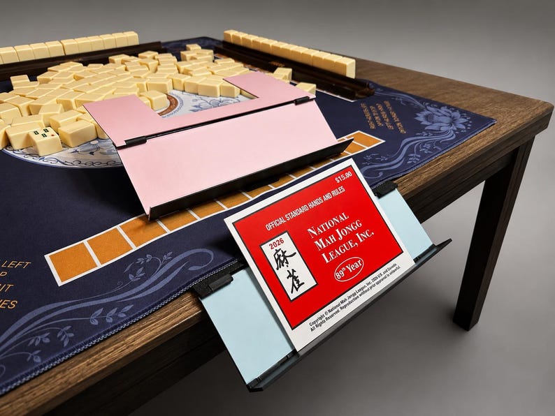 Puede incluir: Un juego de Mahjong completo sobre una mesa de madera. El juego incluye fichas beige, un soporte rosa para fichas y una superficie de juego azul con motivos florales. Tambi&eacute;n se ve un folleto rojo con el texto "National Mah Jongg League, Inc.".
