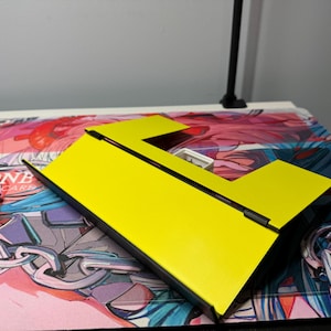 May include: A yellow cardboard box with a black border and a rectangular opening. The box is folded into a triangular shape.