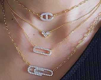 May include: A layered gold necklace with diamond-encrusted pendants. The pendants are shaped like a heart, a rectangle, and a figure eight. The necklace is worn by a person with a dark shirt.