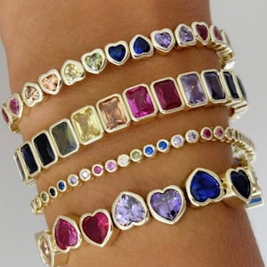 Op de afbeelding: Verschillende gouden armbanden met kleurrijke edelstenen. De armbanden hebben hartvormige, rechthoekige en ronde stenen in verschillende kleuren, waaronder roze, blauw, paars en geel. De armbanden zijn op een pols gestapeld.
