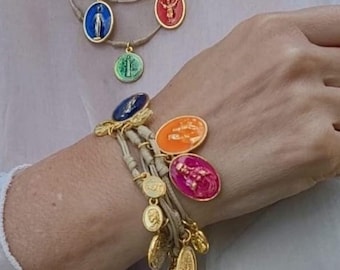 Puede incluir: Un collar de capas con múltiples dijes ovalados en varios colores, incluyendo azul, verde, rojo y naranja. El collar está hecho de cordón beige y presenta una cadena dorada. También se muestra una pulsera a juego con dijes similares.