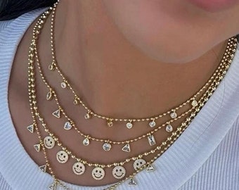 May include: Layered gold chain necklace with a delicate ball chain and multiple charms. The charms include small triangles, round stones, and smiley faces with crystal accents.