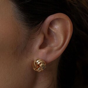 Peut inclure: Gros plan d'une boucle d'oreille à nœud de couleur or. La boucle d'oreille est ronde et présente un motif texturé et entrelacé. La boucle d'oreille est portée sur une oreille.