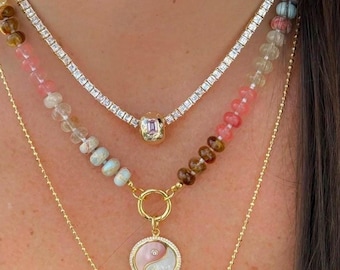 Pode incluir: Um conjunto de colares em camadas com uma corrente dourada, um colar de tênis e um colar de contas com um pingente yin-yang. Os colares exibem uma variedade de cores, incluindo contas rosa, marrons e transparentes, e um pingente dourado com um centro rosa.