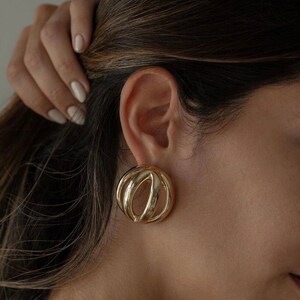 May include: Gold-toned statement earrings with a unique, abstract design. The earrings are circular and feature a series of overlapping, curved lines.