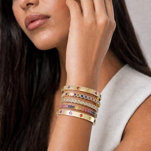 May include: A collection of gold-toned bangles adorns a wrist. Each bangle features colorful gemstones in various designs, including hearts and rows of round stones. The bracelets are stacked, creating a layered look. The gemstones include shades of pink, purple, yellow, and blue.