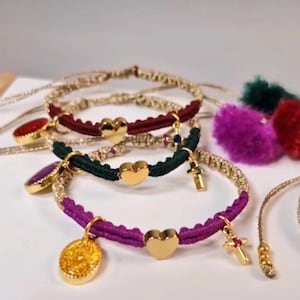 May include: Close-up of several handmade bracelets. The bracelets feature gold-colored accents, including heart-shaped charms and cross pendants. The bands are in various colors, including burgundy, green, and purple. Some bracelets have decorative gemstone charms.