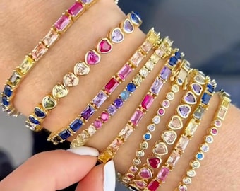 May include: A set of colorful gemstone bracelets with gold chain links. The bracelets feature different colored gemstones, including pink, blue, purple, and yellow. Some bracelets have heart-shaped gemstones, while others have square or round gemstones.