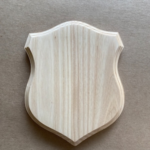 May include: A plain, light wood shield-shaped plaque with a slightly rounded top and bottom. The plaque is made of wood with a natural grain pattern.
