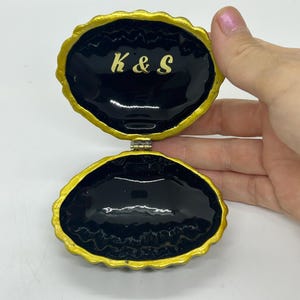 May include: An open clam-shaped trinket box with a glossy black interior and a gold-painted scalloped rim. The upper interior lid features the gold-coloured text K & S. The box is hinged at the centre, revealing a smooth, dark, reflective surface.