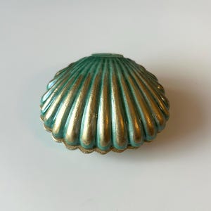 May include: A teal green ceramic seashell with gold detailing. The shell has a ribbed texture and a scalloped edge. The gold paint accentuates the ridges, creating a contrast. A decorative item.