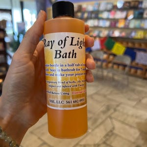 May include: A clear amber glass bottle with a yellow liquid inside. The bottle is labeled "Ray of Light Bath" and has instructions for use. The bottle is being held by a hand.