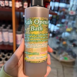 May include: A brown glass bottle with a black cap labeled "Path Opener Bath". The label has green foliage and text that reads "Pour bottle in a half tub of water. Stay in bathtub for 7 minutes and make your prayer request. Proprietary blend of herbs, oils, botanicals and infused with Energy. Well Before Using. External Use Only. SSE, LLC 561 682 0955"