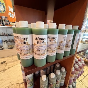 May include: A shelf display of green glass bottles labeled "Money Bath" with instructions for use. The bottles are arranged in rows on a wooden shelf with other bottles of various colors and labels.