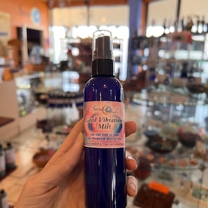 May include: A dark blue spray bottle labeled "Good Vibrations Mist" with a black spray nozzle. The label includes text that reads "Use this mist to raise the vibrations around you." The bottle is held in front of a blurred background.