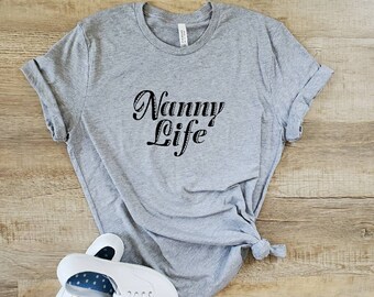 Może przedstawiać: Szary T-shirt z nadrukiem "Nanny Life" w kolorze czarnym na przodzie. Koszulka jest złożona i leży na drewnianej powierzchni. Obok koszulki znajdują się dwa białe buty sportowe z niebieskimi kropkami wewnątrz.