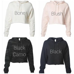 May include: Four cropped hoodies in different colors: bone, blush, black camo, and black. Each hoodie has a drawstring hood and raw hem. The color names are printed on each hoodie.