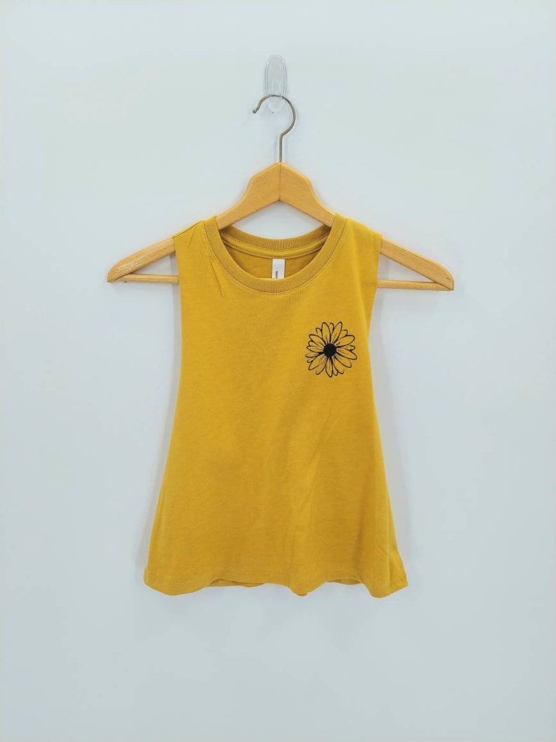 May include: A mustard yellow sleeveless tank top with a black daisy graphic on the front.