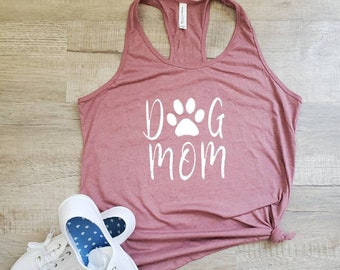 May include: A mauve tank top with a white paw print and the text "Dog Mom" printed on the front. The tank top is tied in a knot at the bottom. A pair of white canvas shoes with blue polka dot lining are on the floor.