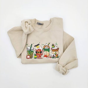 Op de afbeelding: Een beige sweatshirt met een feestelijk ontwerp van vijf verschillende kerst-thema koffie-drankjes. De drankjes zijn versierd met kerstverlichting, snoepstokken en kerstmanmutsen.