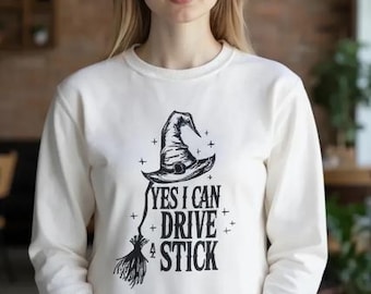 May include: A cream-coloured sweatshirt featuring a black graphic of a witch's hat, broom, and the text "YES I CAN DRIVE A STICK." The design is centered on the front of the long-sleeved top. The sweatshirt is worn by a person.