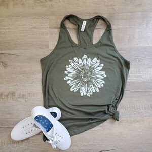 May include: A green tank top with a white daisy graphic. The tank top is made of a soft, comfortable material and has a racerback style. The tank top is paired with white sneakers.
