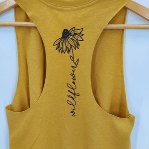 May include: A mustard yellow sleeveless tank top with a black graphic of a wildflower and the text "wildflower" on the back.