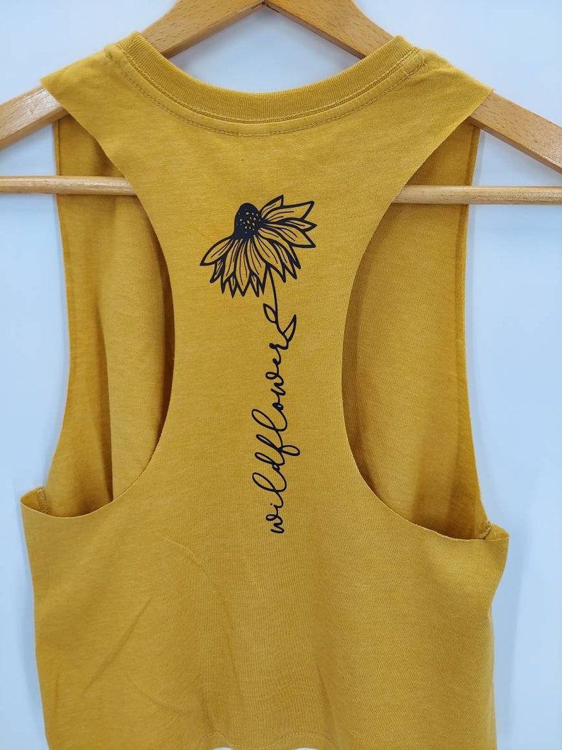 May include: A mustard yellow sleeveless tank top with a black graphic of a wildflower and the text "wildflower" on the back.