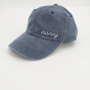 K&ouml;nnte beinhalten: Eine blaue Baseballkappe aus Denim mit dem Wort "nanny" in wei&szlig; auf der Vorderseite gestickt.