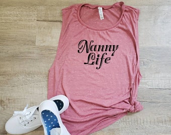 Może przedstawiać: Różowa koszulka bez rękawów z napisem "Nanny Life" w czarnym, ozdobnym pismem. Koszulka leży na drewnianej podłodze, obok pary białych butów z płótna.
