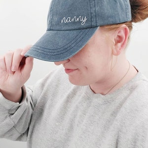 K&ouml;nnte beinhalten: Eine blaue Baseballkappe aus Denim mit wei&szlig;er Stickerei, auf der "nanny" steht.