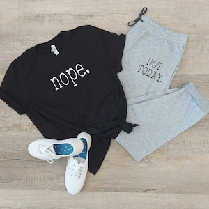 Puede incluir: Una camiseta negra con la palabra "nope" impresa en blanco en la parte delantera, combinada con pantalones de ch&aacute;ndal grises con el texto "NOT TODAY" impreso en negro en la pierna. El conjunto est&aacute; colocado sobre un suelo de madera.