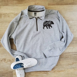 May include: Gray sweatshirt with a black bear silhouette and the word "nanny" inside the bear. The bear has floral details. The sweatshirt has a zipper up the front.