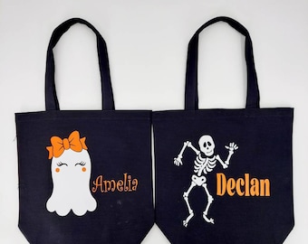 May include: Two black canvas tote bags with Halloween designs. One bag features a white ghost with an orange bow and the name "Amelia". The other bag features a white skeleton with the name "Declan".