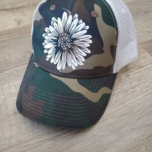 May include: A camouflage trucker hat with a white daisy design. The hat has a mesh back and a brown brim.