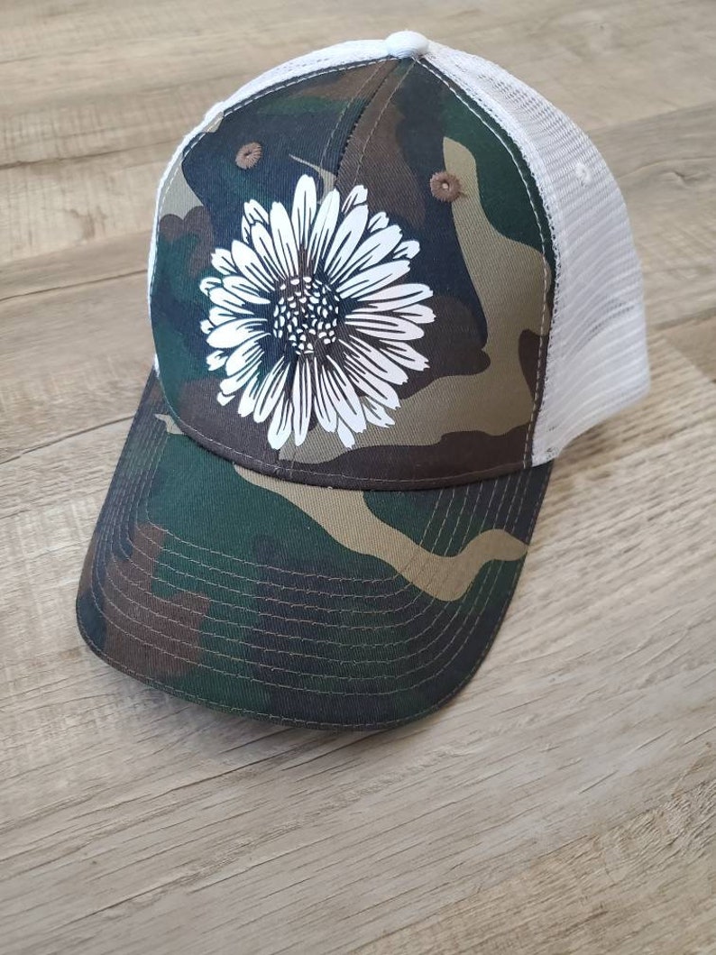 May include: A camouflage trucker hat with a white daisy design. The hat has a mesh back and a brown brim.