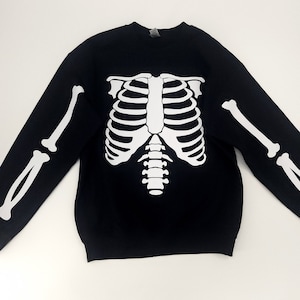 Puede incluir: Sudadera negra con un gráfico de esqueleto blanco impreso en la parte delantera. El gráfico de esqueleto incluye la caja torácica, la columna vertebral y los huesos de los brazos.
