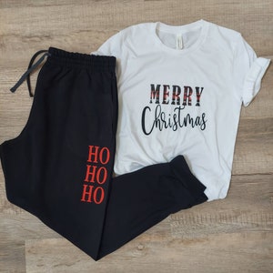 Op de afbeelding: Zwarte joggingbroek met "HO HO HO" in rood op het linkerbeen en een wit T-shirt met "MERRY Christmas" in zwart-rood geruite letters.