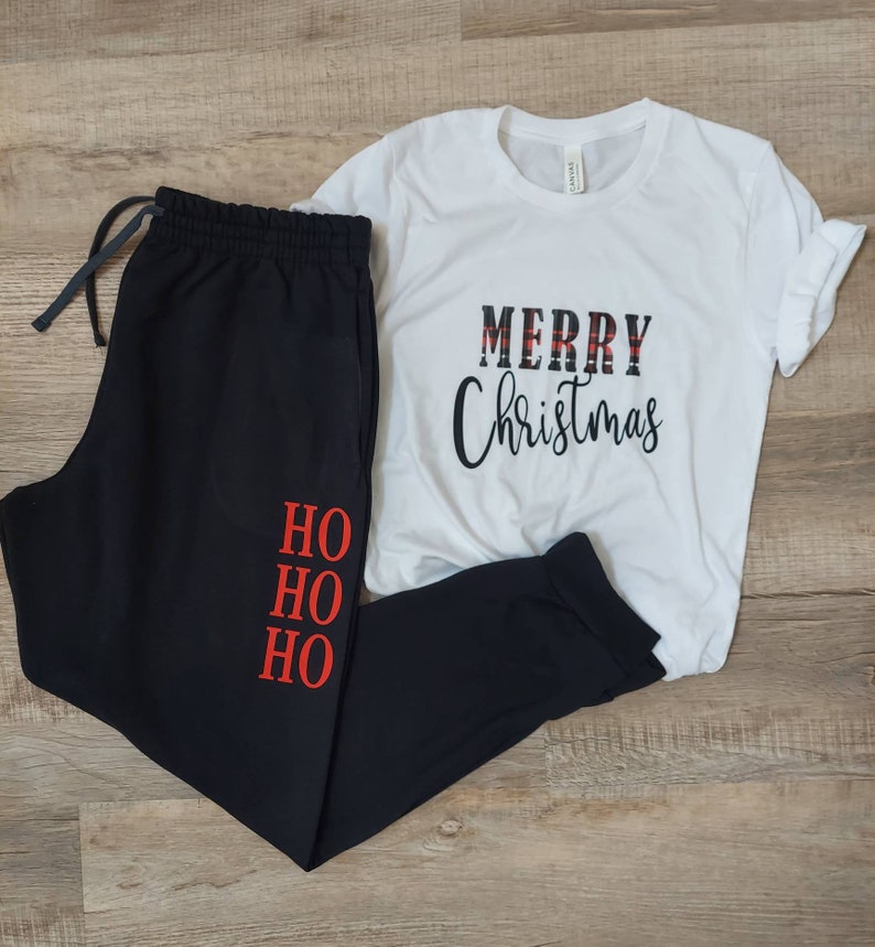 Op de afbeelding: Zwarte joggingbroek met "HO HO HO" in rood op het linkerbeen en een wit T-shirt met "MERRY Christmas" in zwart-rood geruite letters.