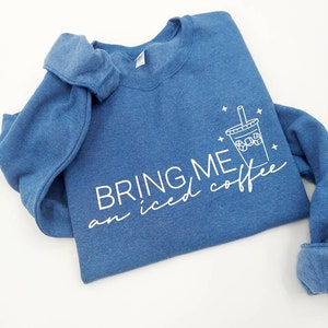 Könnte beinhalten: Ein blaues Sweatshirt mit dem Text "BRING ME an iced coffee" und einer weißen Umrandung eines Bechers mit Eiskaffee und einem Strohhalm.