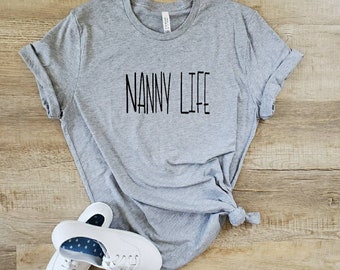 Może przedstawiać: Szara koszulka z nadrukiem "NANNY LIFE" w kolorze czarnym na przodzie. Koszulka leży na drewnianej powierzchni z parą białych butów sportowych.