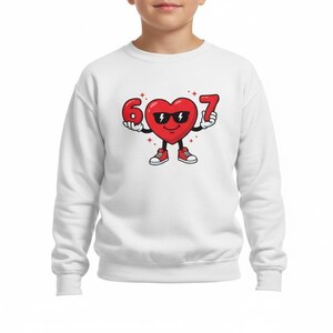 May include: White sweatshirt featuring a cartoon heart character wearing sunglasses and holding the numbers 6 and 7. The heart has arms, legs, and red sneakers. The design is centered on the chest.