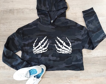 May include: A cropped black and gray camouflage hoodie with a white skeleton hand graphic. The hoodie has a hood and long sleeves. White canvas sneakers with blue polka dot insoles are at the bottom.