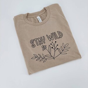 May include: A tan t-shirt with the text "Stay Wild" printed in black. The text is surrounded by a simple floral design.