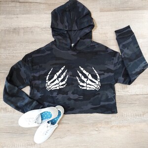May include: A cropped black and gray camouflage hoodie with a white skeleton hand graphic. The hoodie has a hood and long sleeves. White canvas sneakers with blue polka dot insoles are at the bottom.
