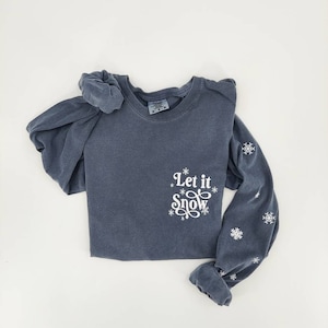 May include: A blue long-sleeved t-shirt with a white "Let it Snow" graphic and snowflake designs on the sleeve.