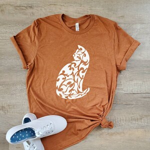 May include: A rust-colored t-shirt with a white silhouette of a cat with a floral design. The shirt is knotted at the bottom and is laying on a wood surface.