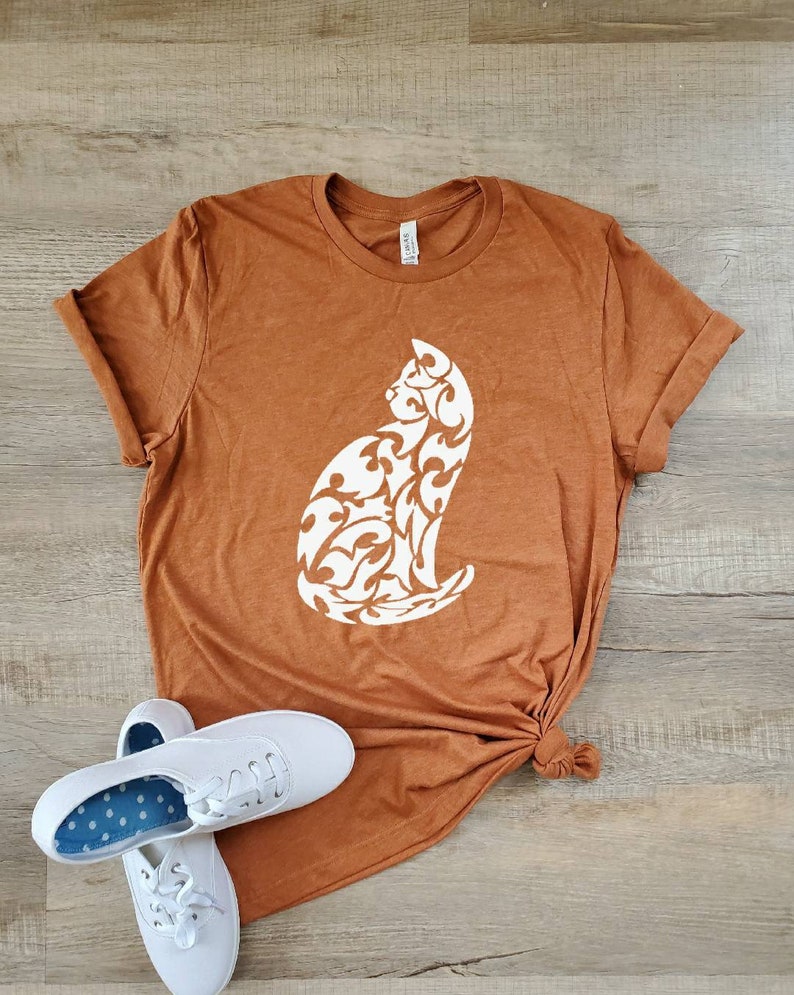 May include: A rust-colored t-shirt with a white silhouette of a cat with a floral design. The shirt is knotted at the bottom and is laying on a wood surface.