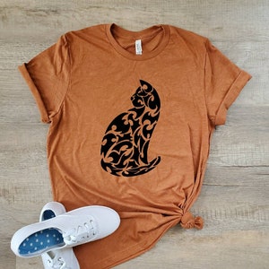May include: A rust-colored t-shirt with a black tribal cat design. The shirt is made of a soft, comfortable material and has a relaxed fit.